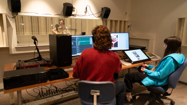Kaksi opiskelijaa istuu äänitysstudiossa ja tarkkailee äänityslaitteistoa. Kolmas opiskelija soittaa kitaraa studion lasin takana.
